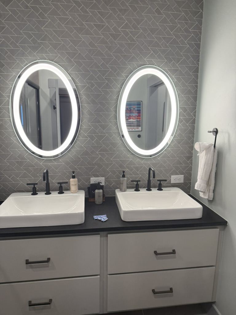 Double vanity with two vessel sinks, black faucets, and oval LED mirrors on a grey chevron tile wall.